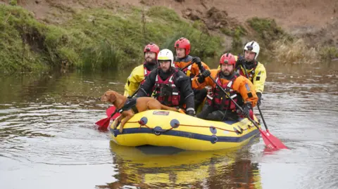 PA Media Five search and rescue team members crossing  the River Aln near Alnwick, Northumberland, on Wednesday in a yellow boat. A dog in the boat is looking out.