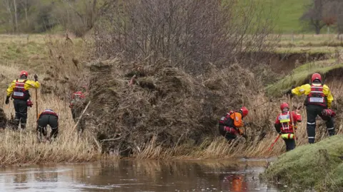 PA Several people wearing waterproof gear and lifejackets and helmets are scouring the river bank. There is debris on the bank.